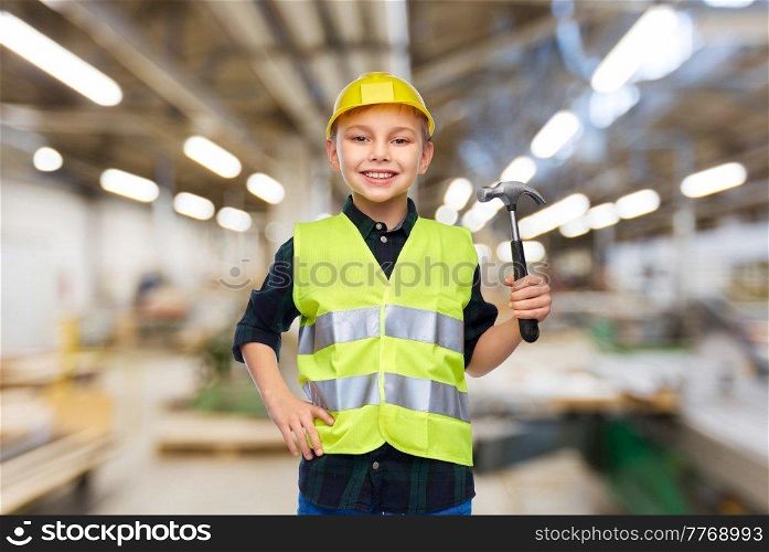 industry, manufacture and profession concept - happy smiling little boy in protective helmet and safety vest with hammer over workshop background. boy in protective helmet and vest with hammer