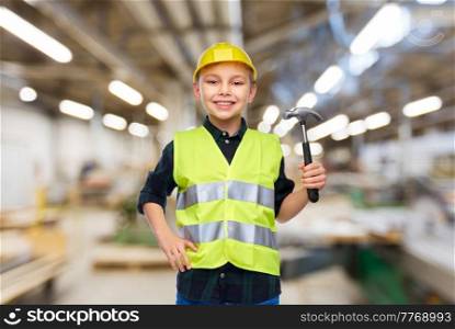 industry, manufacture and profession concept - happy smiling little boy in protective helmet and safety vest with hammer over workshop background. boy in protective helmet and vest with hammer