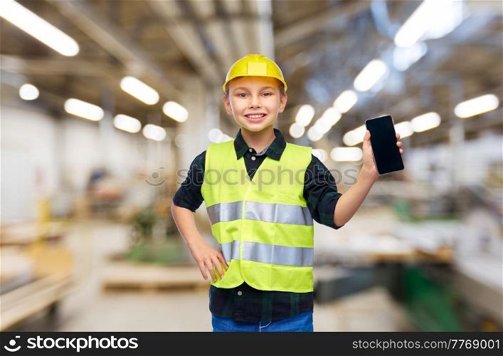 industry, manufacture and profession concept - happy smiling little boy in protective helmet and safety vest with smartphone over workshop background. little boy in helmet and safety vest with phone