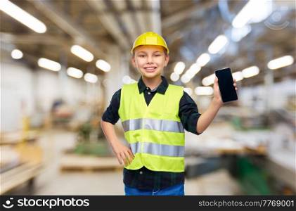 industry, manufacture and profession concept - happy smiling little boy in protective helmet and safety vest with smartphone over workshop background. little boy in helmet and safety vest with phone