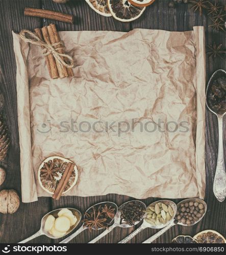 Ingredients in iron spoons for preparation of mulled wine drink on a brown wooden background, in the middle mashed brown kraft paper for writing a recipe, top view