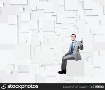 Innovative technologies. Young handsome businessman sitting on white cube holding mobile phone in hand