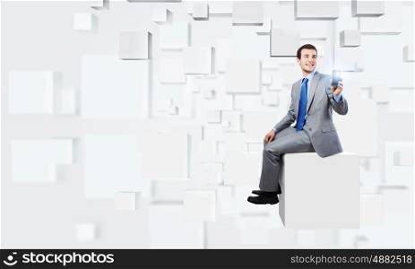 Innovative technologies. Young handsome businessman sitting on white cube holding mobile phone in hand