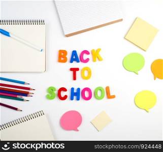 inscription back to school from multi-colored plastic letters and a stack of notebooks with blank white sheets on a white background, top view
