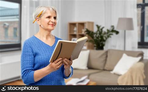 inspiration, memoirs and old people concept - portrait of smiling senior woman in blue sweater with diary or notebook and pencil over home living room background. senior woman with pencil and diary or notebook