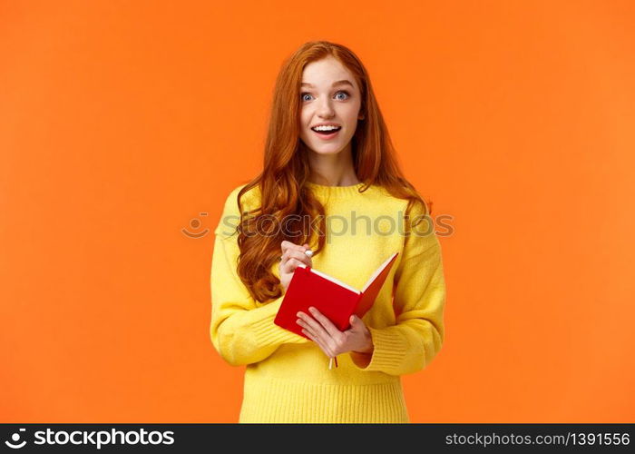 Inspired and upbeat, dreamy fascinated redhead girl writing down interesting lecture, smiling and gazing with admiration or enthusiasm, fill-in notebook schedule, standing orange background.. Inspired and upbeat, dreamy fascinated redhead girl writing down interesting lecture, smiling and gazing with admiration or enthusiasm, fill-in notebook schedule, standing orange background