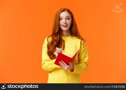 Inspired and upbeat, dreamy fascinated redhead girl writing down interesting lecture, smiling and gazing with admiration or enthusiasm, fill-in notebook schedule, standing orange background.. Inspired and upbeat, dreamy fascinated redhead girl writing down interesting lecture, smiling and gazing with admiration or enthusiasm, fill-in notebook schedule, standing orange background