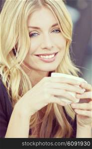 Instagram filter style beautiful smiling young woman with blond hair and blue eyes drinking coffee or tea from a white cup
