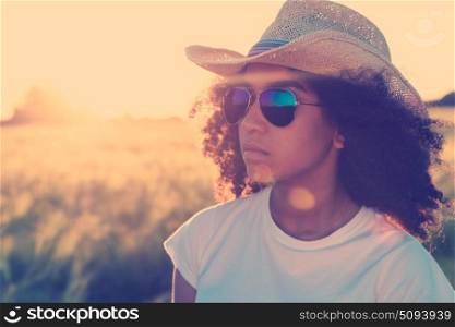 Instagram filter style photo of beautiful happy mixed race African American female girl teenager young woman wearing reflective aviator sunglasses and cowboy hat in a cornfield at golden sunset or sunrise