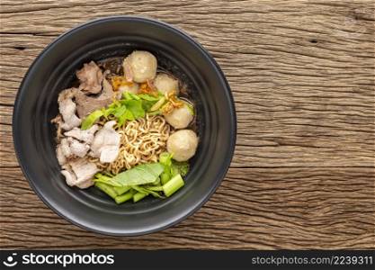instant noodles soup with beef, meatball, pork liver, water spinach, morning glory, celery, fried garlic and herb in black ceramic bowl on rustic wood texture background, top view, flat lay