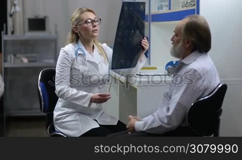 Intellectual female healthcare employee looking at elderly male patient&acute;s MRI scan in hospital office. Beautiful female doctor in eyeglasses and lab coat holding tomography and pointing at it during medical checkup.