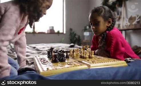 Intellegent mixed race sisiters spending leisure in domestic interior playing chess on bed. Adorable pensive children capturing chess figures during chess match at home. Side view. Close up. Dolly shot. Slow motion.