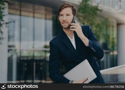 Intelligent businessman speaks on phone during walk to office waits for business meeting to start wears formal clothing uses modern gadgets for communication poses outdoor near blurred background. Intelligent businessman speaks on phone during walk to office