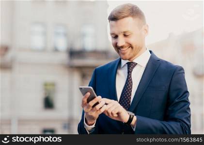 Intelligent businessman with positive expression, uses application on smart phone for navigating in big city, has working trip, wears elegant clothes, has happy look. People and occupation concept
