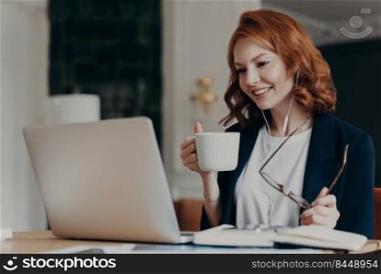 Intelligent female student has online course, concentrated in screen of laptop computer, learns foreign languages distantly, drinks tea or coffee, poses at desktop. Woman worker has video conference
