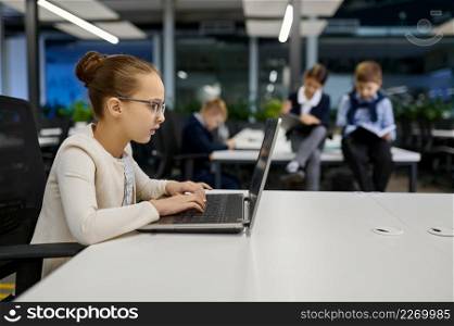 Intelligent little girl child working on laptop in office. Children coworker team on background. Girl child working on laptop in office