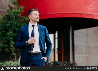 Intelligent male banker dressed in formal outfit, keeps hand in pocket, looks thoughtfully aside, tries to plan his working day, poses outdoor, stands near building. People, business and lifestyle
