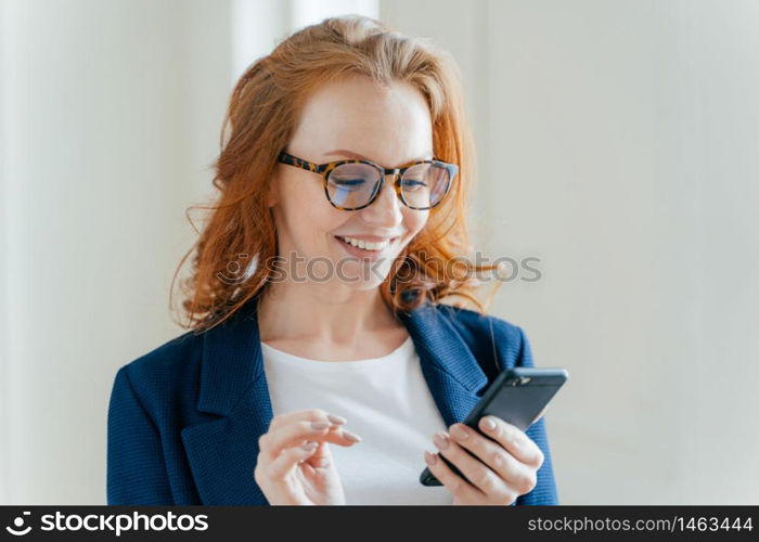 Intelligent positive female entrepreneur in elegant apparel, happy to recieve message on mobile phone, finds out about raising profits, has red hair, smiles happily, poses indoor in modern office