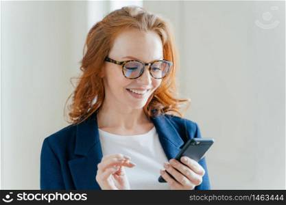 Intelligent positive female entrepreneur in elegant apparel, happy to recieve message on mobile phone, finds out about raising profits, has red hair, smiles happily, poses indoor in modern office