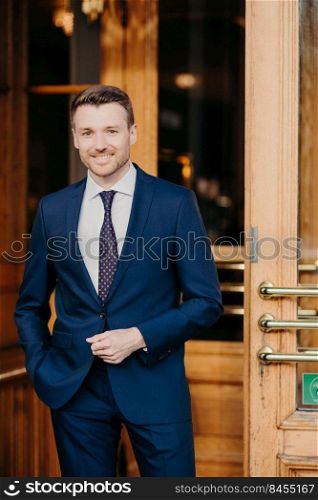 Intelligent young male director or boss wears black suit, has happy expression, confident in his success, stands near doors of comapny, being pleased to meet with business partners. Man employee