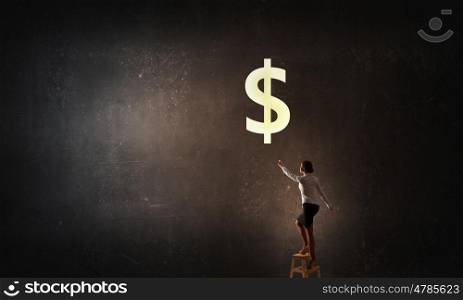 Intention to become rich. Young attractive businesswoman standing on chair and reaching money bag