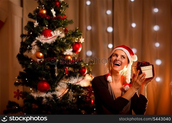 Interested woman near Christmas tree shaking present box trying to guess what&rsquo;s inside&#xA;