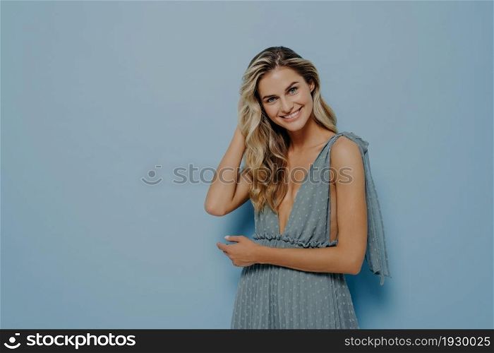 Interested young blonde woman in romantic blue dress gently touching her hair, smiling with tilted head, expressing positive emotions while standing isolated in front of blue background. Blonde woman in blue dress touching her hair