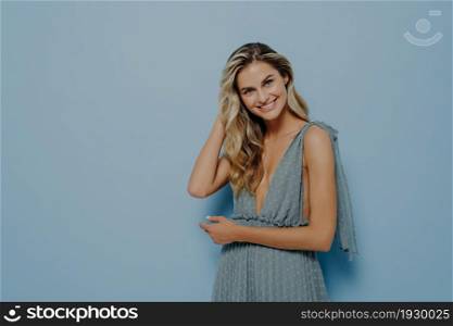 Interested young blonde woman in romantic blue dress gently touching her hair, smiling with tilted head, expressing positive emotions while standing isolated in front of blue background. Blonde woman in blue dress touching her hair