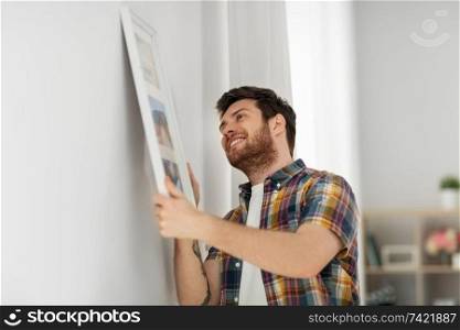 interior decoration and renovation concept - smiling man hanging picture in frame to wall at home. man hanging picture in frame to wall at home