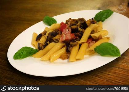 International Italian food with a close-up of pasta penne rigatoni with basil, ham and aubergines on wooden table.