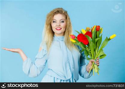 International women day, eight march. Lovely woman blonde girl with red yellow tulips, elegant dress holding open empty hand, copy space for product. On blue. woman with tulips bunch, open hand