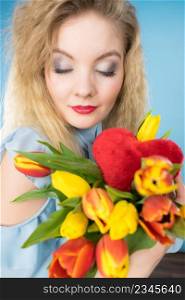 International womens or valentines day. Attractive happy woman blonde hair fashion make up holding tulips bunch and red heart sign. On blue. Woman holds tulips and red heart