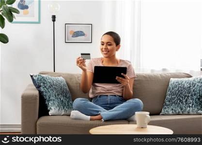 internet banking, online shopping and technology concept - happy smiling african american woman sitting on sofa with tablet pc computer and credit card at home. happy woman with tablet pc and credit card at home