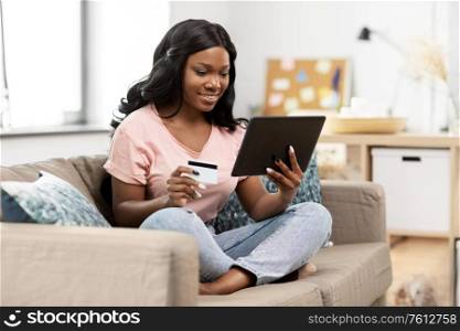 internet banking, online shopping and technology concept - happy smiling african american woman sitting on sofa with tablet pc computer and credit card at home. happy woman with tablet pc and credit card at home