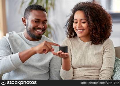 internet of things and technology concept - happy african american couple with smart speaker sitting on sofa at home. african american couple with smart speaker at home