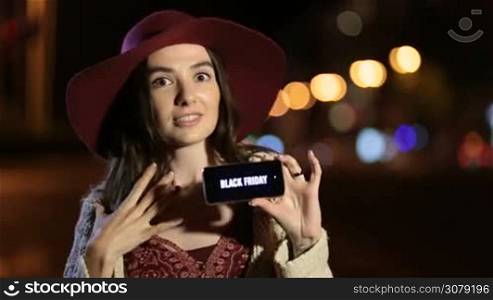 Internet shopping woman holding smartphone with black friday sale, advertising text on screen over colorful bokeh of night city streetlights background. Fashionable female showing mobile phone with black friday sign on screen on night city street.