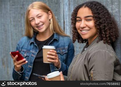 Interracial friends pretty blonde teenager and mixed race African American girl female young woman using their smart phones for social media and drinking takeout coffee outside