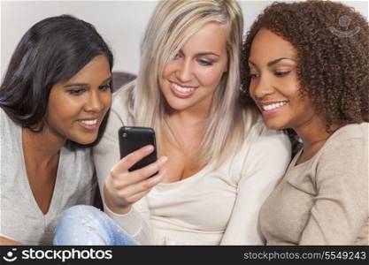 Interracial group of three beautiful young women girl friends at home sitting together on a sofa smiling and having fun using cell phone smartphone