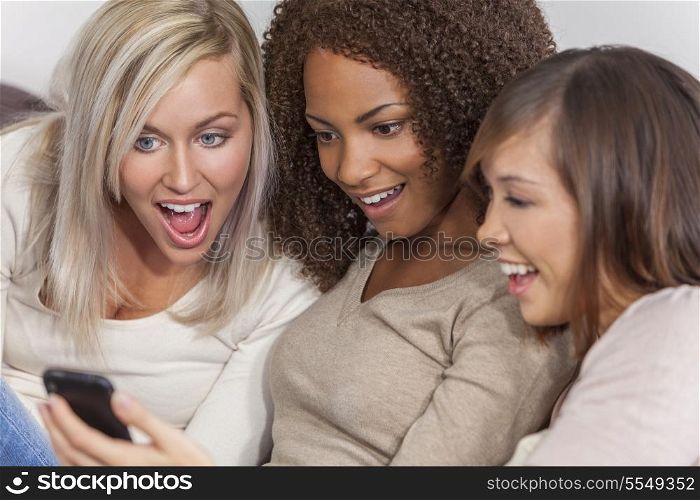 Interracial group of three beautiful young women girl friends at home sitting together on a sofa smiling, surprised and shocked using cell phone smartphone