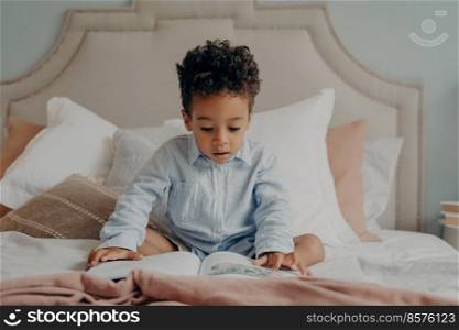 Intrigued cute afro american boy preschool child with curly hair reading book or exploring pictures inside while sitting comfortably on bed, making surprised expression. Children development concept. Intrigued afro american preschool child looking at colorful pictures inside of book while sitting on big bed