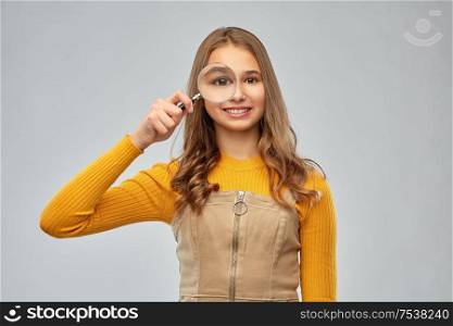 investigation, discovery and vision concept - happy teenage girl looking through magnifying glass over grey background. teenage girl looking through magnifying glass