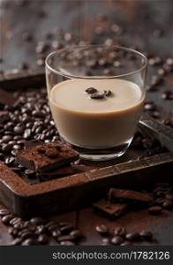 Irish cream baileys liqueur in glass in wooden tray with coffee beans and powder with dark chocolate on dark wood background. Macro