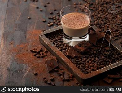 Irish cream baileys liqueur in glass with coffee beans and powder with dark chocolate in wooden tray on dark wood background. Space for text