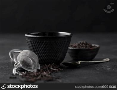 Iron cup with black tea and strainer infuser with loose tea leaf on black with silver spoon. Macro