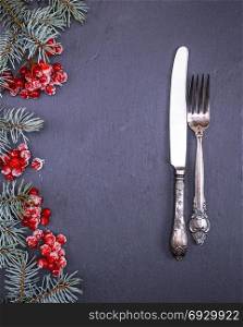 iron vintage fork and knife on a black background, decorated with a branch of spruce and clusters of viburnum, empty space in the middle
