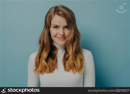 Irresistible beauty. Portrait of gorgeous young smiling girl with nice natural make-up wearing white long sleeved turtleneck posing isolated over blue studio wall background with empty space. Gorgeous young smiling woman with nice natural make-up posing isolated over blue studio background