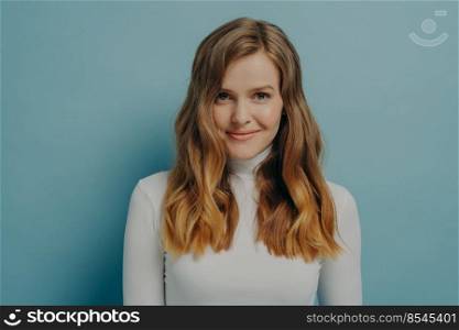 Irresistible beauty. Portrait of gorgeous young smiling girl with nice natural make-up wearing white long sleeved turtleneck posing isolated over blue studio wall background with empty space. Gorgeous young smiling woman with nice natural make-up posing isolated over blue studio background