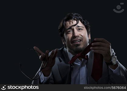 Irritated young businessman holding glasses and tie over black background