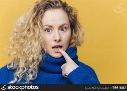 Isolated shot of amazed beautiful female with curly blonde hair, keeps fore finger on lower lip, listens someone attentively, dressed in blue sweater, isolated over yellow background. Emotions
