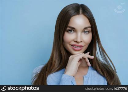 Isolated shot of attractive brunette woman with calm expression, healthy skin and makeup, keeps hand under chin, cares about complexion, looks beautiful, isolated on blue background, models in studio